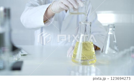 A scientist, wearing a lab coat and white protective gloves, is pouring a yellow oily liquid from one beaker to another in laboratory, close up. Medicine and science concept A scientist, wearing a lab coat and white protective gloves, is pouring a yellow oily liquid from one beaker to another in laboratory, close up. Medicine and science concept 123694273