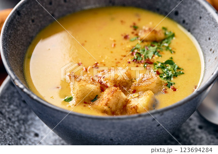Bowl of mashed potato soup with a spoon on the plate. The bowl is filled with yellow soup puree and topped with croutons, parsley and ground red pepper. Bowl of mashed potato soup with a spoon on the plate. The bowl is filled with yellow soup puree and topped with croutons, parsley and ground red pepper. 123694284