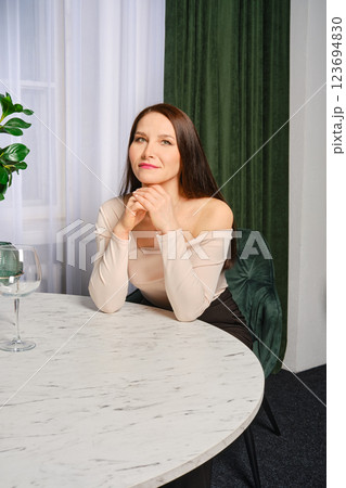 Middle-aged woman resting her chin on hands at a round table in a stylish living room Middle-aged woman resting her chin on hands at a round table in a stylish living room 123694830