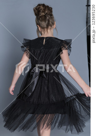 A young girl in a black tulle dress twirls gracefully, showing the elegant back details of her outfit. This image captures the beauty of movement in children's formal fashion, highlighting the dress's 123695230