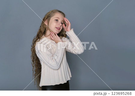 A young girl with long, wavy hair gently touches her face while wearing a cozy sweater, showcasing her natural beauty. This image highlights the importance of children's hair care and promotes 123695268