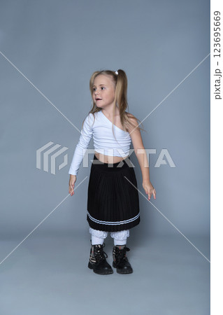 A young girl with blonde pigtails dressed in a white one-shoulder top and pleated black skirt stands confidently. The playful contrast between her trendy outfit and innocent demeanor highlights 123695669