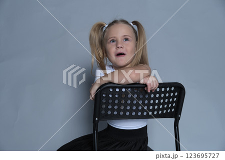 Smiling girl with pigtails leaning on a black chair, exuding confidence and comfort in her own skin, posing for a portrait. 123695727