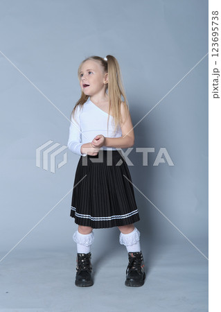 young girl showcases a chic look in a one-shoulder white top paired with a black pleated skirt and black boots. Her confident stance and playful expression bring out the modern trends in children's 123695738