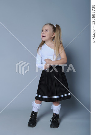 young girl with blonde pigtails is standing confidently against a grey background, wearing a stylish one-shoulder white top, a black pleated skirt, white lace socks, and black boots. Her pose is young girl with blonde pigtails is standing confidently against a grey background, wearing a stylish one-shoulder white top, a black pleated skirt, white lace socks, and black boots. Her pose is 123695739