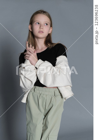 A young girl with long blonde hair, dressed in a black top and light green pants, stands thoughtfully with her hands clasped. This image conveys a sense of contemplation and innocence, reflecting 123696238