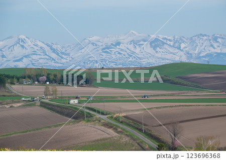 春の丘陵地帯の農作業と残雪の山並み　十勝岳連峰 123696368