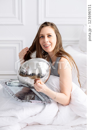 Young woman in braces and pajamas sits on bed among white sheets and opens tray. Morning breakfast in bed at hotel Young woman in braces and pajamas sits on bed among white sheets and opens tray. Morning breakfast in bed at hotel 123696551