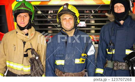 Portrait of young happy fireguards in protective uniforms against background of fire truck at station. Male firefighters in full equipment looking into camera with positive emotions near big red car Portrait of young happy fireguards in protective uniforms against background of fire truck at station. Male firefighters in full equipment looking into camera with positive emotions near big red car 123707268