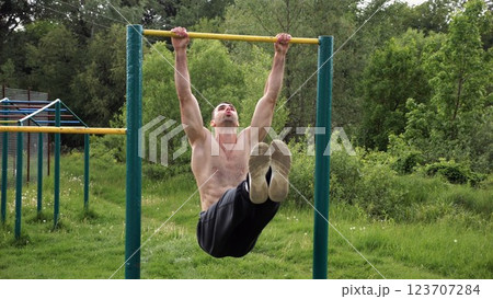 Athletic man trains abdominal muscles raising legs on horizontal bar at sports ground. Young sportsman exercising on pull-up bar outdoor. Sporty athlete working out at nature. Healthy lifestyle Athletic man trains abdominal muscles raising legs on horizontal bar at sports ground. Young sportsman exercising on pull-up bar outdoor. Sporty athlete working out at nature. Healthy lifestyle 123707284
