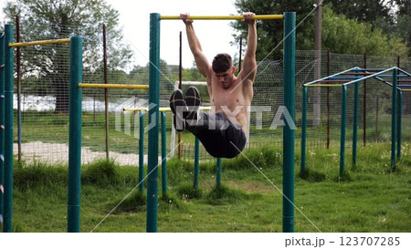 Athletic man trains abdominal muscles raising legs on horizontal bar at sports ground. Young sportsman exercising on pull-up bar outdoor. Sporty athlete working out at nature. Healthy lifestyle Athletic man trains abdominal muscles raising legs on horizontal bar at sports ground. Young sportsman exercising on pull-up bar outdoor. Sporty athlete working out at nature. Healthy lifestyle 123707285