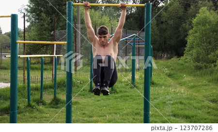 Strong guy training abdominal muscles on horizontal bar at sports ground. Young sportsman exercising on pull-up bar outdoor. Sporty athlete working out at nature. Concept of sportive lifestyle Strong guy training abdominal muscles on horizontal bar at sports ground. Young sportsman exercising on pull-up bar outdoor. Sporty athlete working out at nature. Concept of sportive lifestyle 123707286