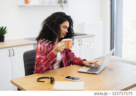 African woman early 30s working on laptop, drinking coffee, seated in kitchen workspace 123717175