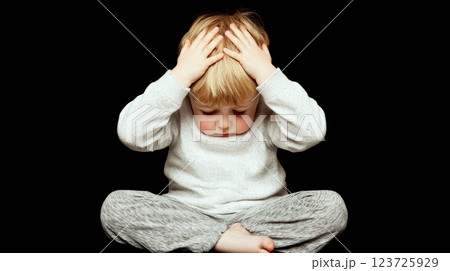 A child sits cross-legged in a dark setting, holding their head with hands. Concept of contemplation and childhood emotions. 123725929