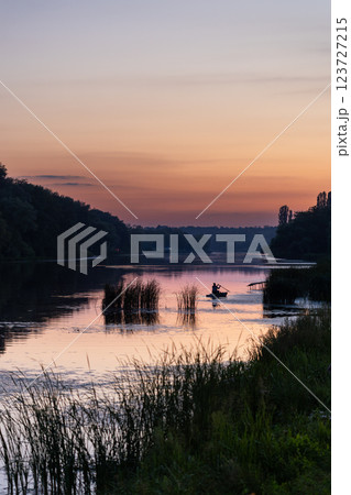 A lone fisherman rows a small boat on a calm river at sunset. The sky glows in shades of orange and purple, reflecting on the water. Tall grasses and trees frame the peaceful scene. A lone fisherman rows a small boat on a calm river at sunset. The sky glows in shades of orange and purple, reflecting on the water. Tall grasses and trees frame the peaceful scene. 123727215