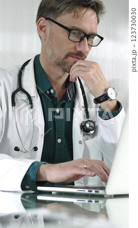 A male doctor in a green shirt under a medical coat is thinking while using technology in healthcare. Man is typing on a laptop on a glass desk in the clinic, vertical portrait view. Medicine concept 123730030
