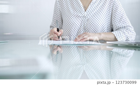 Businesswoman in white dotted blouse is signing a contract at a sleek glass desk in a modern office, showcasing professionalism and focus in her work environment. Business people concept Businesswoman in white dotted blouse is signing a contract at a sleek glass desk in a modern office, showcasing professionalism and focus in her work environment. Business people concept 123730099