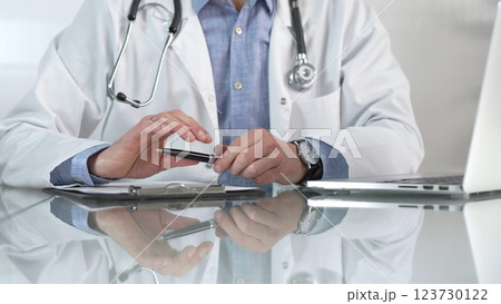 Male doctor wearing a blue shirt, stethoscope and lab coat is gesturing with hands while explaining a diagnosis to a patient across a glass desk with a laptop. Medicine concept 123730122