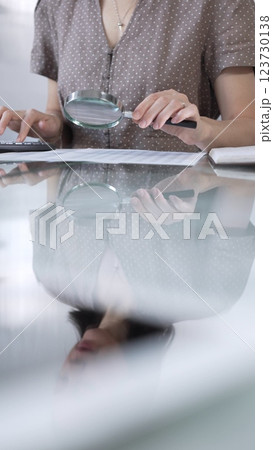 Businesswoman accountant in brawn dotted blouse reviewing financial reports with magnifying glass and calculator in office, vertical view. Taxes, audit in business 123730138