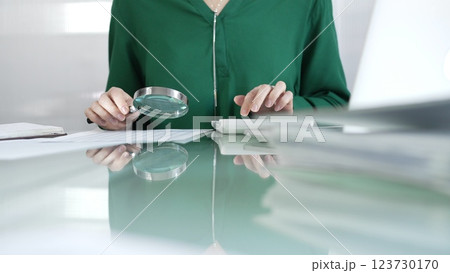 Businesswoman in green blouse carefully reviewing financial reports with magnifying glass and calculator, conducting audit and analyzing data in office. Taxes, audit in business Businesswoman in green blouse carefully reviewing financial reports with magnifying glass and calculator, conducting audit and analyzing data in office. Taxes, audit in business 123730170