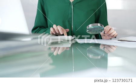 Businesswoman in green blouse carefully reviewing financial reports with magnifying glass and calculator, conducting audit and analyzing data in office. Taxes, audit in business 123730171