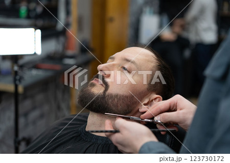 Skilled barber grooming clients beard using professional scissors, delivering precise styling within contemporary barbershop setting 123730172