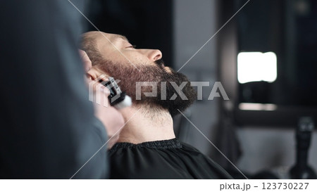 Professional barber trimming client's beard with electric razor, delivering precise styling within contemporary grooming salon setting 123730227