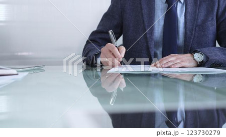 Businessman in a professional attire signing a contract with a pen on a modern glass desk in a contemporary office environment. Business people or audit concept Businessman in a professional attire signing a contract with a pen on a modern glass desk in a contemporary office environment. Business people or audit concept 123730279