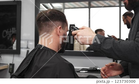 Skilled barber trimming client's hair using electric razor, creating contemporary hairstyle in modern barbershop setting with precise, professional technique 123730300