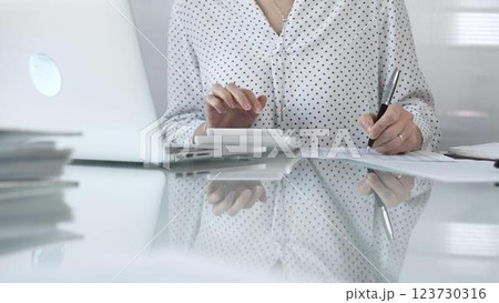 Unknown businesswoman using a calculator to crunch numbers and jotting down meticulous notes at a sleek glass desk in a contemporary office, closeup view. Taxes, audit in business 123730316
