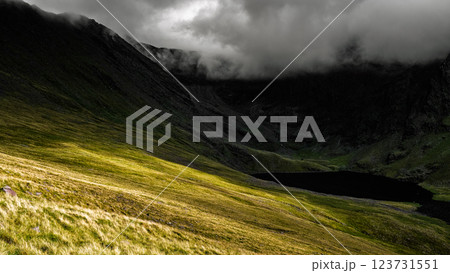 Lush green hills contrast with dark peaks shrouded in clouds, while a tranquil lake lies quietly below, embodying the wild beauty of Ireland's landscape. Lush green hills contrast with dark peaks shrouded in clouds, while a tranquil lake lies quietly below, embodying the wild beauty of Ireland's landscape. 123731551
