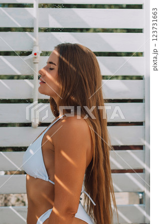A woman with long brown hair is standing in front of a white wall. She is wearing a white bikini and she is enjoying the sun A woman with long brown hair is standing in front of a white wall. She is wearing a white bikini and she is enjoying the sun 123731663