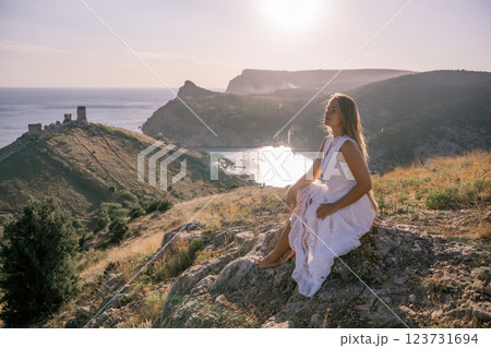 A woman in a white dress sits on a rock overlooking a body of water. The scene is serene and peaceful, with the woman enjoying the view and the calmness of the surroundings. 123731694