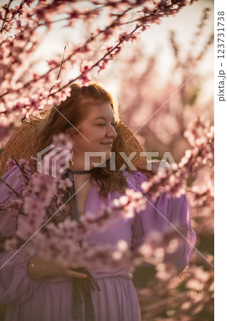 Woman blooming peach orchard. Against the backdrop of a picturesque peach orchard, a woman in a long pink dress and hat enjoys a peaceful walk in the park, surrounded by the beauty of nature. 123731738