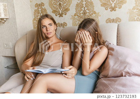 mom and daughter sitting on a couch, one of them reading a book. The other woman is looking away, possibly in a state of shock or disbelief. Concept of tension and unease. 123732193