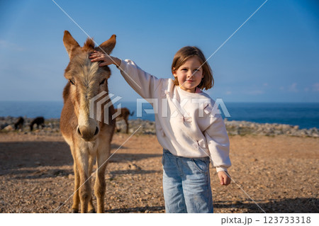 A little girl gently pets a donkey in the National Donkey Park on the northeasternmost cape of Northern Cyprus. Heartwarming connection between children and wildlife in a remote natural setting 123733318