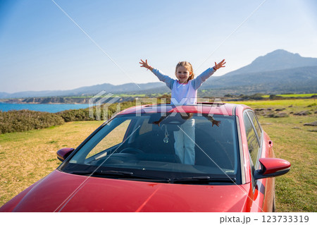 Little girl joyfully sticks out of car sunroof with raised hands against the backdrop of mountains in Northern Cyprus. The image symbolizes freedom, adventure, and the excitement of travel with child Little girl joyfully sticks out of car sunroof with raised hands against the backdrop of mountains in Northern Cyprus. The image symbolizes freedom, adventure, and the excitement of travel with child 123733319