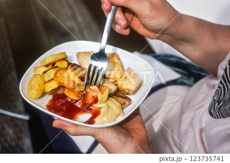 Woman hand holding fork and plate with grilled potatoes, and tofu (soy bean curd) with ketchup and mustard - vegetarian barbecue / grilling 123735741