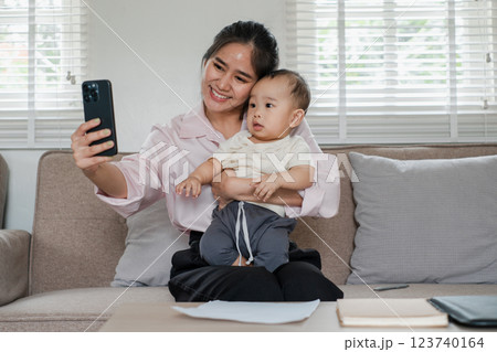 Smiling mother holding her baby while taking a selfie on a cozy couch in a well-lit living room. 123740164