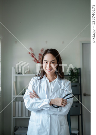 Smiling female doctor in a white coat holding a stethoscope, standing in a modern medical office with a bookshelf and plants. Smiling female doctor in a white coat holding a stethoscope, standing in a modern medical office with a bookshelf and plants. 123740175