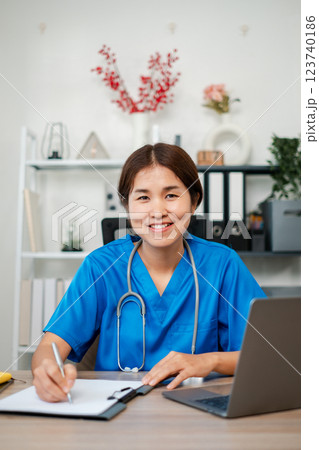 Friendly healthcare worker in blue scrubs writing on clipboard at desk with laptop, showcasing a professional medical environment. Friendly healthcare worker in blue scrubs writing on clipboard at desk with laptop, showcasing a professional medical environment. 123740186