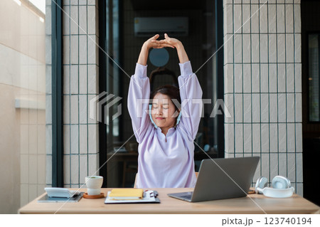 Woman in casual attire stretches at a desk with a laptop and coffee, promoting relaxation and productivity in a modern home office. Woman in casual attire stretches at a desk with a laptop and coffee, promoting relaxation and productivity in a modern home office. 123740194