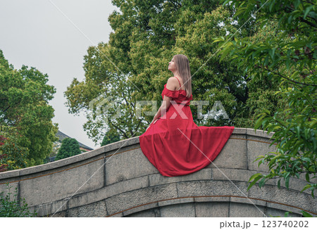 Woman in a flowing red dress on a stone bridge in a tranquil garden during cloudy weather Woman in a flowing red dress on a stone bridge in a tranquil garden during cloudy weather 123740202