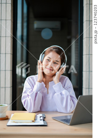Smiling woman wearing headphones, listening to music, working at home with a laptop and coffee on the desk. Smiling woman wearing headphones, listening to music, working at home with a laptop and coffee on the desk. 123740203