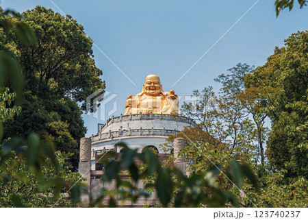 A magnificent golden Buddha statue sits atop a temple, surrounded by vibrant trees and foliage under a bright blue sky. 123740237