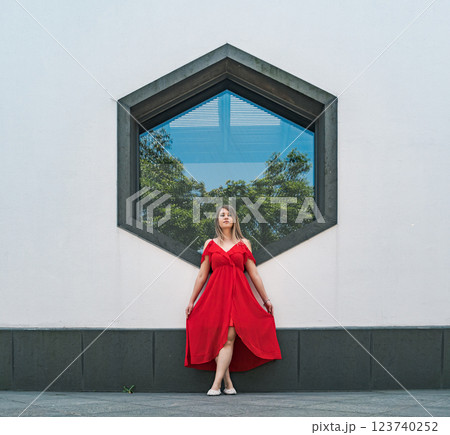 A woman stands elegantly in a flowing red dress, striking a pose in front of a unique hexagonal window. 123740252