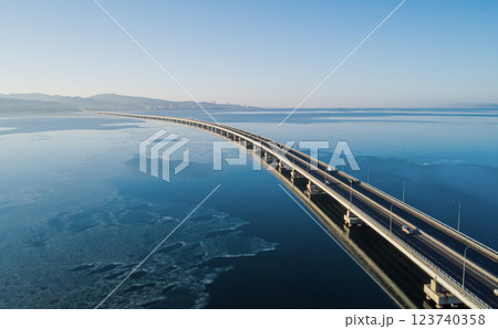 Aerial view of a long road bridge stretching over calm waters at dawn with distant mountains and city skyline 123740358