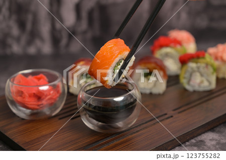Chopsticks holding sushi roll and dipping in soy sauce. Set of sushi roll with rice and fish on wooden serving board. Dark black texture background. Close up. Japan menu. Asian food 123755282
