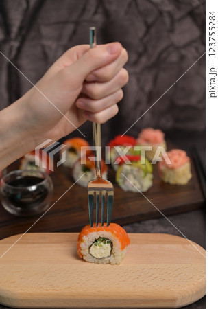 Woman's hand prick sushi roll with a fork. Set of sushi roll with rice and fish on wooden serving board. Dark black texture background. Close up. Japan menu. Asian food 123755284