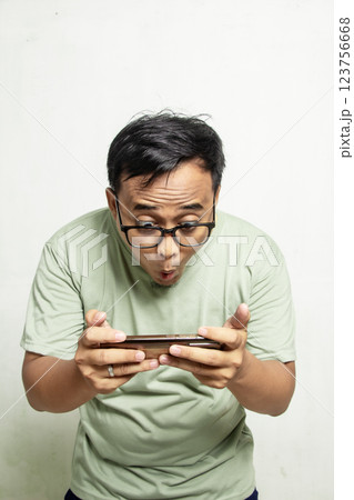 Portrait of an Asian Indonesian man with glasses who is surprised while playing a mobile game on his smartphone or news about something fun. Half body man and sage green shirt Portrait of an Asian Indonesian man with glasses who is surprised while playing a mobile game on his smartphone or news about something fun. Half body man and sage green shirt 123756668